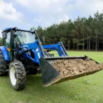 A New Holland tractor with a front-end loader bucket full of fill dirt being moved across a residential yard for a square foot garden project.