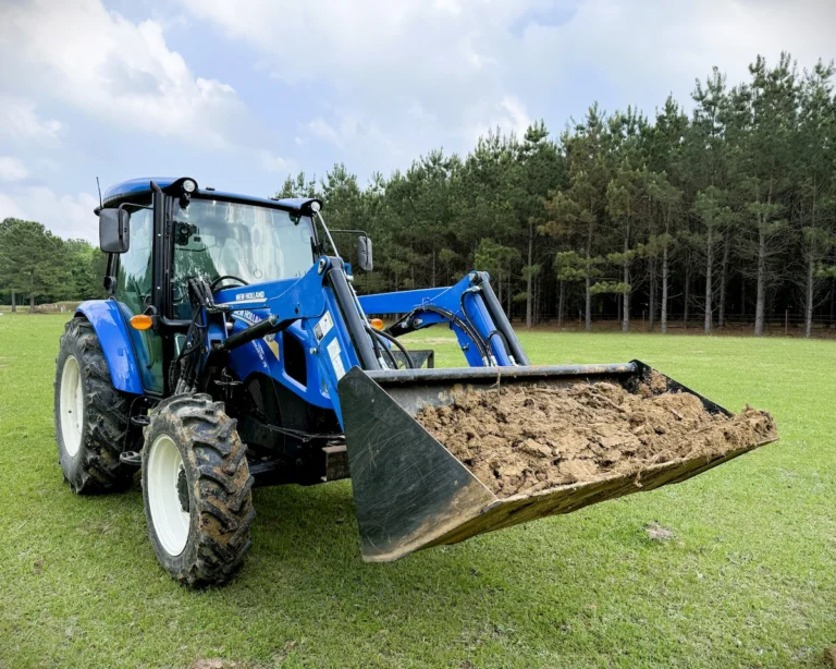 A New Holland tractor with a front-end loader bucket full of fill dirt being moved across a residential yard for a square foot garden project.